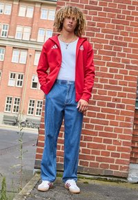 Giacca rossa Adidas, canottiera bianca, pantaloni in denim blu larghi e sneakers bianche con righe rosse. Riccioli testurizzati completano il look.