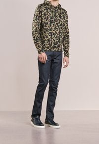 Felpa con cappuccio mimetica in tonalità verde e marrone, abbinata a jeans blu scuro. Modello indossa scarpe da ginnastica nere con suole bianche, in piedi su una superficie chiara.