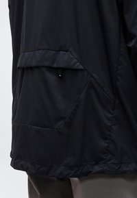 Κοντινό πλάνο πίσω τσέπης μαύρου μπουφάν με καπάκι και μικρό ιμάντα έλξης, φορεμένο πάνω από γκρίζο παντελόνι.