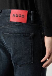Jean en denim bleu foncé avec un patch en cuir rouge portant l'inscription "HUGO" à la ceinture, doté de poches arrière standard et de détails de couture visibles.