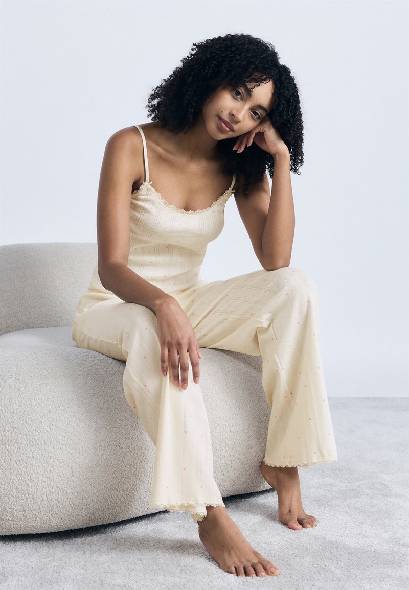 Νεαρή γυναίκα με σγουρά μαλλιά, φορώντας κρεμ πιτζάμες, καθισμένη ξυπόλητη σε μια υφασμάτινη μπεζ καρέκλα μπροστά σε φωτεινό φόντο.