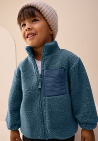 Veste en polaire couleur sarcelle avec fermeture éclair à l'avant, col montant et une poche en tissu bleu marine. Texture douce, coupe décontractée et manches longues.