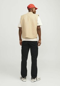 Jachetă bej fără mâneci peste un tricou alb, asortată cu pantaloni cargo negri și adidași gri. Căciula este roșie, iar fundalul este neutru.