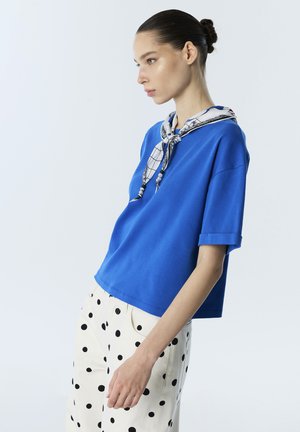 Jonge vrouw met haar in een knot, die een blauw topje met korte mouwen draagt, een zwart-witte polkadot broek en een bedrukte sjaal om haar nek geknoopt