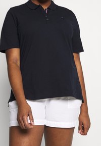 Personne portant un polo noir à manches courtes et un short blanc, se tenant avec les bras détendus devant un fond uni.