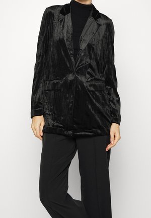 Personne portant un blazer en velours noir à un bouton, un col roulé noir et un pantalon noir, sur un fond clair uni.