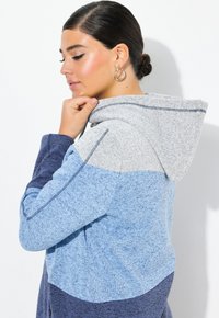 Mikina s kapucí s barevným blokovým designem v odstínech světle modré, šedé a námořnické. Měkká, strukturovaná tkanina s kontrastními švy.