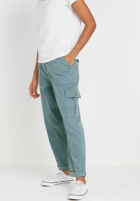 Lichtgroene cargobroek met een relaxte pasvorm, voorzien van zijzakken en opgerolde zomen, gecombineerd met een witte V-hals t-shirt en witte sneakers.