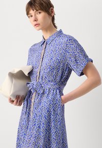Vestido azul com padrão, de mangas curtas, gola e cinto na cintura. O tecido apresenta um design floral em tons de creme e laranja. Segura uma mala bege.