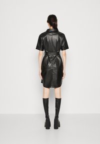 Zwarte faux leren overhemdjurk met korte mouwen, een ceintuur in de taille, knie-lengte en zijsplitten. Model gecombineerd met zwarte laarzen tot de knie.