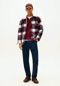 Jauns vīrietis, ģērbies sarkanā raibā žaketē, tumši sarkanā krekliņā, tumši zilās biksēs, ar brūnu cepuri un staigušanas apaviem, stāv ar vienu roku kabatā.