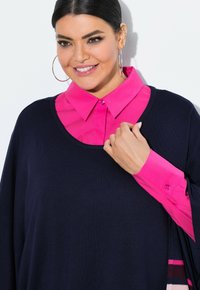 Marineblaues Strickoberteil, das über einem knallpinken Hemd mit Kragen und Knopfdetails getragen wird. Das Oberteil hat eine lockere Passform und einen runden Ausschnitt.