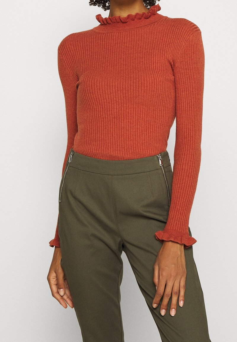 Pomarańczowa bluzka w prążki z wysokim, falbaniastym kołnierzem i dopasowanymi długimi rękawami; zestawiona z oliwkowymi spodniami z zamkami błyskawicznymi w kieszeniach.