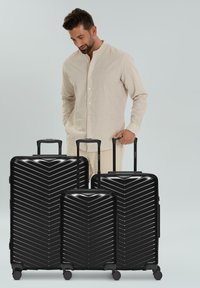 Homme en tenue beige se tenant derrière trois valises rigides noires de tailles variées avec des poignées extensibles, sur un fond gris clair.
