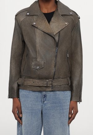 Person iført en mørkebrun læderbikerjakke med lynlåsede lommer, bælte i taljen og lyseblå denimjeans.