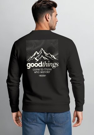 Mann trägt schwarzen Sweatshirt mit Bergmotiv und Text "good things come to those who wander" auf dem Rücken, steht vor einfarbigem Hintergrund.