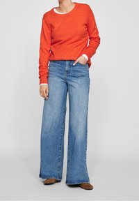 Pull en maille rouge avec un col rond, associé à un jean en denim bleu clair à jambes larges. Le jean a un ourlet brut et un léger effet usé. Chaussures marron.