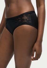 Gros plan sur une personne portant des sous-vêtements noirs avec bordure en dentelle, montrant les hanches, le bas-ventre et la main droite sur un fond uni.