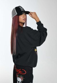 Černá oversized mikina s vyšitými grafikami, spárovaná s černou a šedou kšiltovkou s detailním designem. Dlouhé, rovné vlasy jsou viditelné.