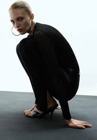 Top negro de manga larga con patrón texturizado, combinado con pantalones negros ajustados y mules de tacón abierto, colocados sobre una superficie oscura.