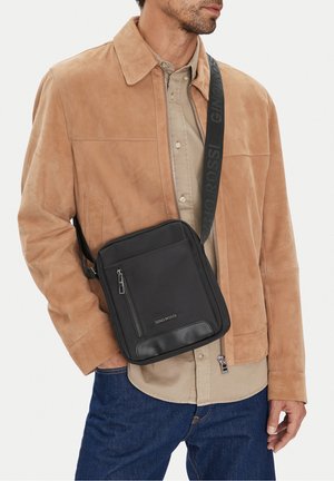 Uomo che indossa una giacca in camoscio color caffelatte, camicia beige e jeans blu con una borsa nera a tracolla con etichetta "GINO ROSSI" sulla cinghia della spalla.