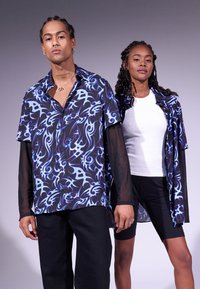 Una maglietta nera a maniche corte con motivi astratti blu, indossata sopra una maglietta bianca. Pantaloni corti neri e pantaloni neri con texture completano il look.