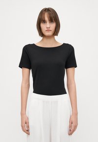 Camiseta preta de manga curta com decote redondo. Feita de tecido liso, com silhueta ajustada. Usada com calças brancas pregueadas.