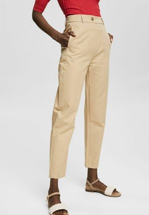 Pantalons en coton beige à taille haute, avec poches latérales et fermeture à bouton. Portés avec des sandales en cuir tressé beige.