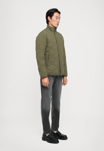 Jachetă cu acoladă de culoare verde măslin, cu guler înalt, închidere zip și buzunare laterale, combinată cu blugi din denim gri și pantofi negri.
