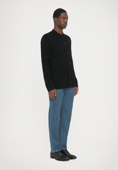 Zwarte button-up cardigan met een kraag, gemaakt van een gestructureerde stof. Gecombineerd met blauwe denim broek en zwarte oxford schoenen.
