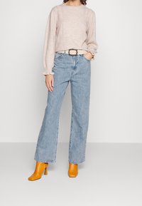 Ljusbeige tröja med texturerad detalj, högmidjade blå denimjeans och gula klackstövlar. Accessoarer inkluderar ett beige bälte.