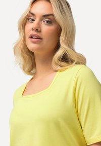 T-shirt in cotone giallo con scollatura quadrata, maniche corte e una texture liscia. Il design presenta dettagli minimi e una vestibilità comoda.