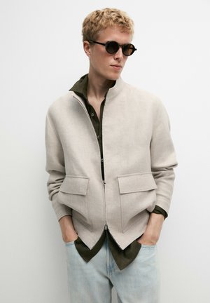 Junge Person mit kurzen blonden Haaren, die runde dunkle Sonnenbrillen trägt, eine hellbeige Jacke mit Reißverschluss über einem dunklen Shirt und hellblaue Jeans, mit Händen in den Taschen.