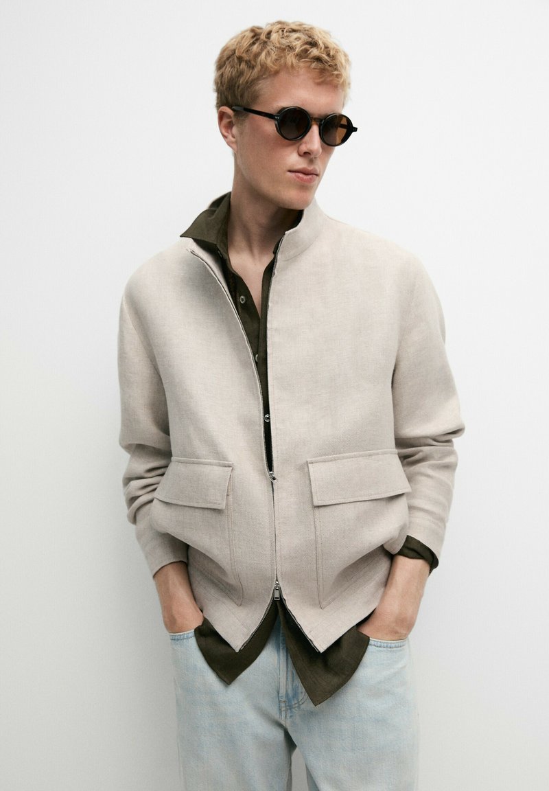 Jeune personne aux cheveux blonds courts portant des lunettes de soleil rondes et foncées, une veste zippée beige clair par-dessus un t-shirt foncé, et un jean bleu clair, les mains dans les poches.