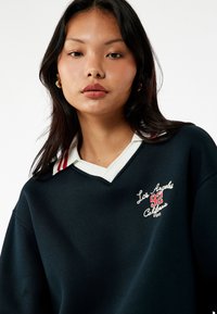 Felpa navy con colletto a strisce bianche e rosse, caratterizzata da testo ricamato: "Los Angeles 92 California" in bianco e rosso.