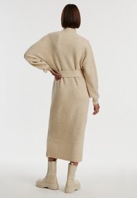 Beige gestrickter langer Cardigan mit hohem Kragen und tailliertem Gürtel, ausgestattet mit langen Ärmeln und einem gerippten Textur, kombiniert mit cremefarbenen Stiefeletten.