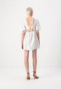 Λευκό φόρεμα με μανίκια μανταρίνι και άνοιγμα στην πλάτη, δεμένο με φιόγκο; κατασκευασμένο από ελαφρύ ύφασμα με υφή; συνδυασμένο με μπεζ πέδιλα με λουράκια.