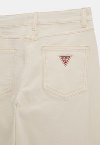 Des jeans en denim crème avec une taille haute, un design à cinq poches et un patch logo triangulaire rouge "Guess" sur la poche arrière.