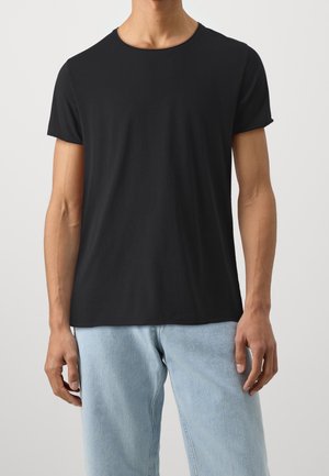 Hombre vestido con una camiseta negra de manga corta y jeans de mezclilla de color azul claro frente a un fondo blanco liso.