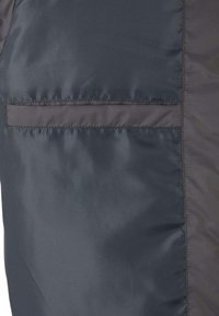 Mørk grå pufferjakke med en glatt, blank tekstur og en horisontal brystlomme. Har stiftede sømmer og et strømlinjeformet design.