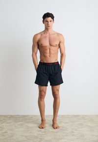 Čierne plavecké šortky s elastickým pásom, na ktorých je červené logo "HUGO". Model stojí naboso na textúrovaných svetlých podlahách.