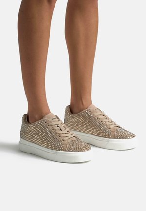 Jambes portant des baskets basses beiges couvertes de petits strass brillants et des semelles en caoutchouc blanches, debout sur une surface blanche unie.