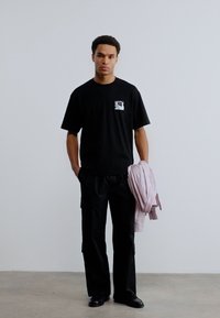 Νέος άνδρας ντυμένος στα μαύρα κρατάει ένα ανοιχτό ροζ σακάκι στο ένα χέρι, μπροστά σε απλό φωτεινό φόντο.