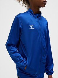 Persona con una chaqueta deportiva azul con cremallera de Hummel, con las manos en los bolsillos, mostrada desde la mitad del torso hasta la barbilla sobre un fondo liso.