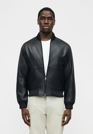 Mand iført en sort læderbomberjakke over en hvid skjorte og lyse beige bukser, stående mod en ensartet lys baggrund.