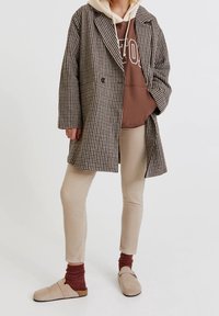 Cappotto oversize a scacchi marrone e crema, indossato sopra un felpa marrone, abbinato a pantaloni chiari e mocassini beige.