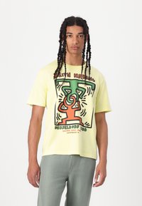 Light yellow cotton t-shirt featuring a graphic print in green, black, and orange, with short sleeves and a relaxed fit.