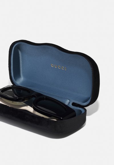 Svarta solglasögon inne i ett svart sammetset med blå inredning. Guld "GUCCI"-logotyp visas inuti. Vinklad ram, mörka linser.
