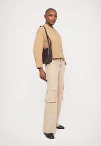 Donna che indossa una giacca trapuntata beige, pantaloni cargo beige, scarpe nere e porta una borsa a tracolla marrone scuro, in piedi su uno sfondo bianco.