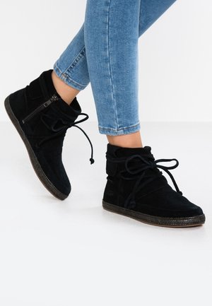 Bottines noires en daim avec fermeture éclair latérale et lacets portées avec un jean bleu retroussé sur fond blanc.